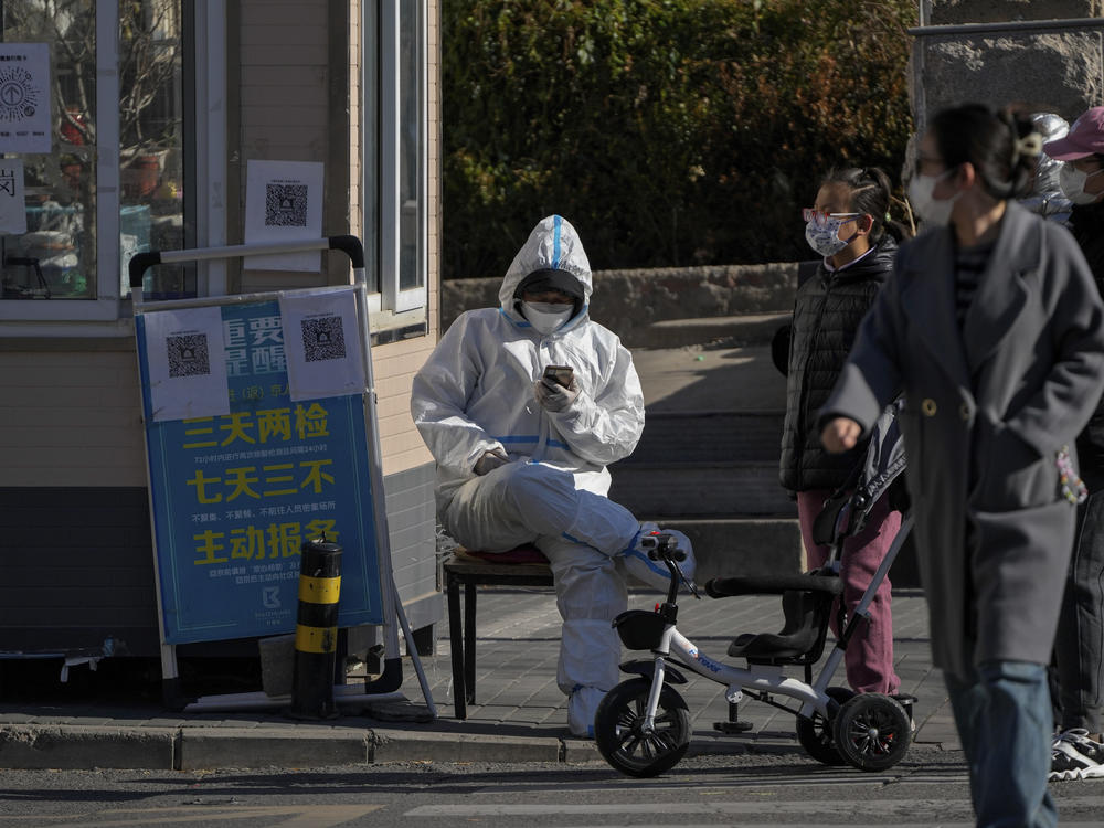 Residents walk by a security guard in protective suit browsing his phone at a main entrance gate to a neighborhood in Beijing, Thursday, Dec. 1, 2022.
