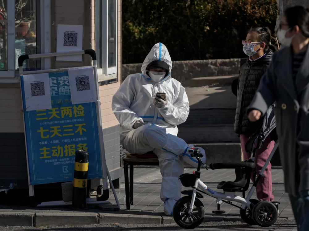 2022 年 12 月 1 日，星期四，北京某小区的主要入口，居民从一名身穿防护服的保安身旁走过，他正在浏览手机。