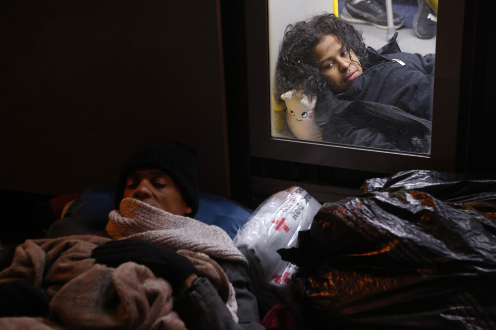 A migrant lays covered in blankets in front of Sacred Heart Church (left) on Wednesday while another lays on the floor of a bus meant to allow people to warm themselves after crossing the Rio Grande.