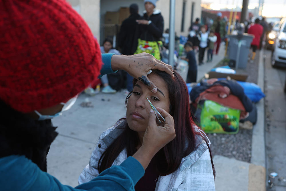 An unsheltered asylum-seeking migrant from Venezuela (right) has her eyebrows done by another  migrant from Colombia on the sidewalk near Sacred Heart Church on Thursday.