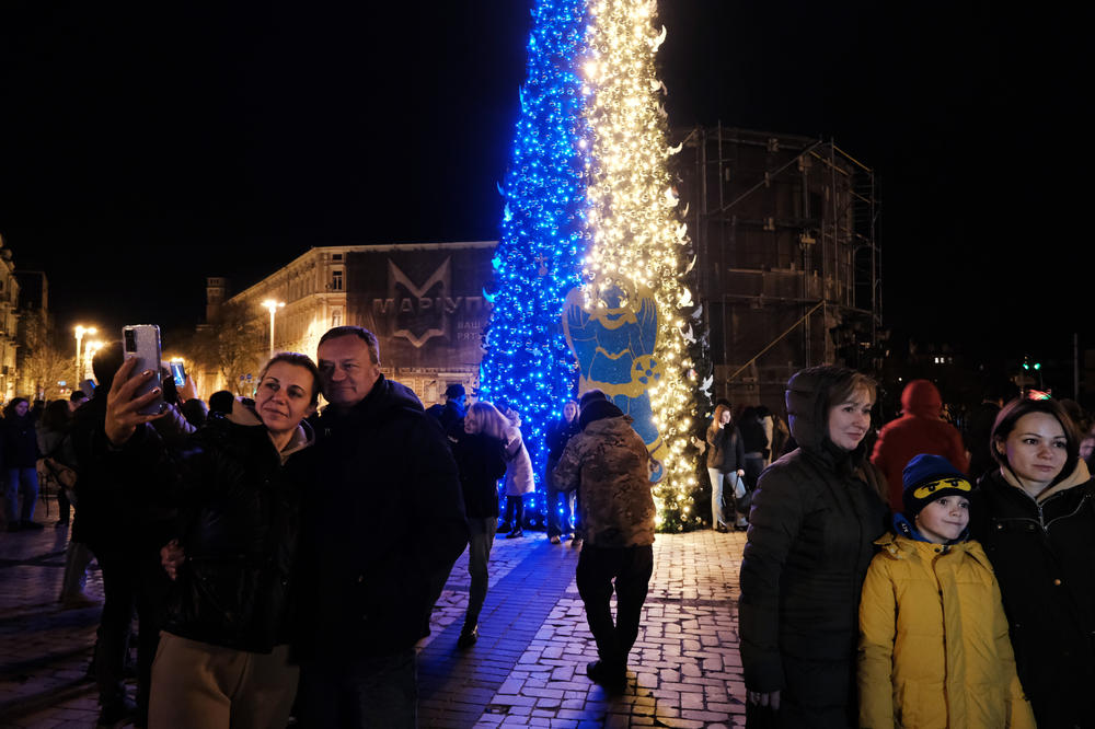 People gather near a Christmas tree decorated in the colors of the Ukrainian flag in a Kyiv square on New Year's Eve. Many residents are without power or water as the Russian missile assault continues throughout the country.