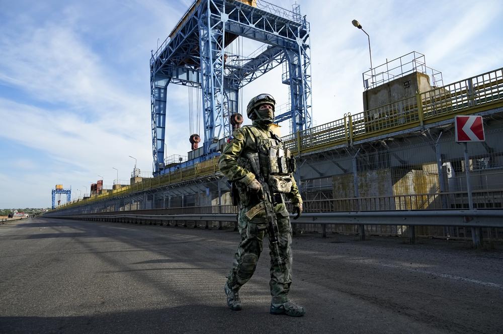 A Russian soldier patrols an area at the Kakhovka Hydroelectric Station on May 20, 2022, in a photograph taken during a trip organized by the Russian Ministry of Defense. The blue gantry cranes, which control locks along the dam, can be seen in the background.