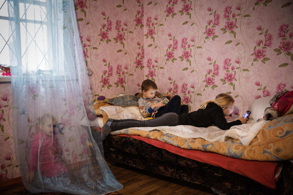 Miroslava Grinik, 8, and her younger brother Kirill, 5, on their smart phones at their new home in Poltava region, which they moved into in October 2022. Their cousin Alisa, plays on the floor by the bed. December 2022.
