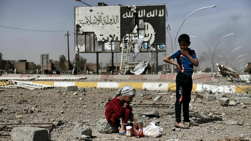 Iraqi children sit amid the rubble of a street in Mosul's Nablus neighborhood in front of a billboard bearing the logo of the Islamic State group on March 12, 2017.
