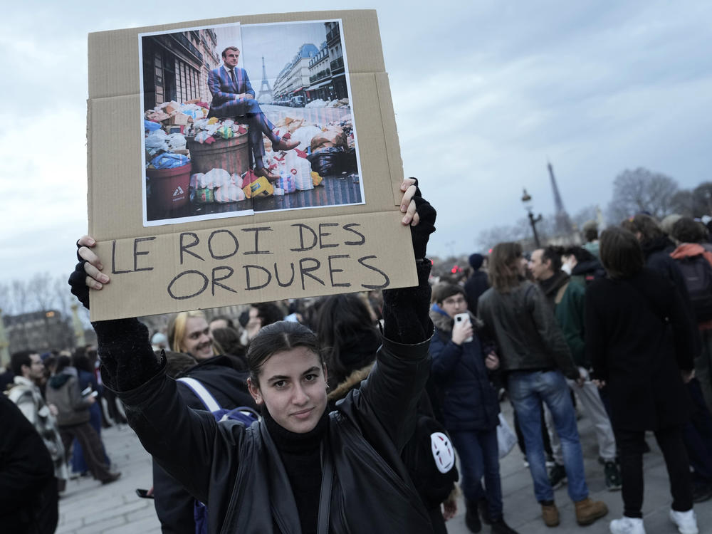 A woman holds a placard depicting French President Emmanuel Macron sitting on garbage cans that reads, 