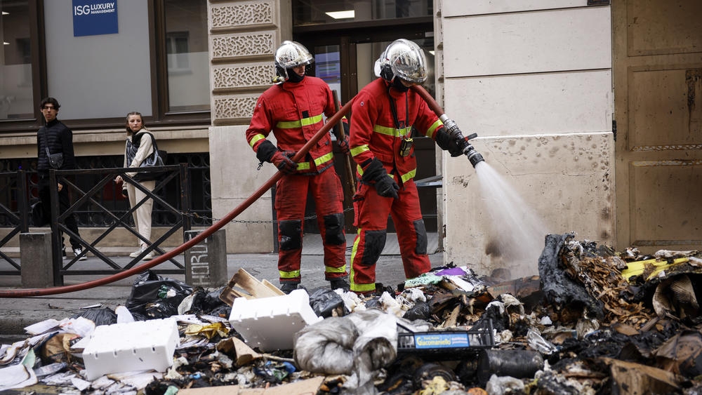 Firemen controlling the remains of a garbage fire from Thursday night protests against the retirement bill in Paris on Friday. French President Macron's office says a state visit by Britain's King Charles III is postponed amid mass strikes and protests.