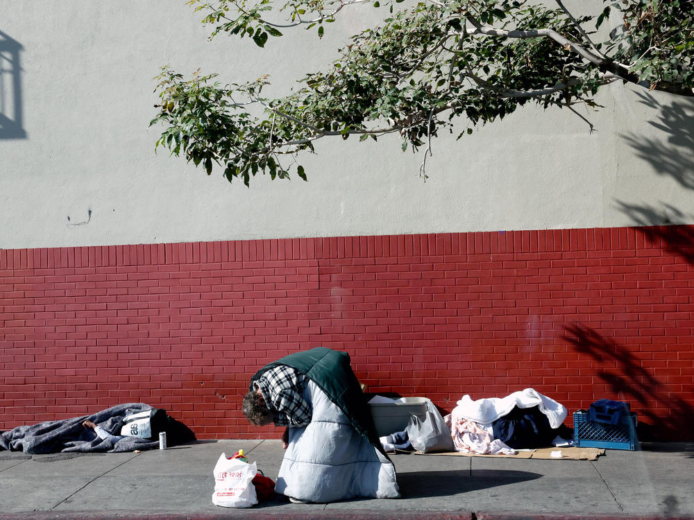 A person kneels on the sidewalk as another person sleeps in the Skid Row community on Dec. 14, 2022 in Los Angeles, California. The Skid Row community is home to thousands of people who either live on the streets or in tents or makeshift shelters.