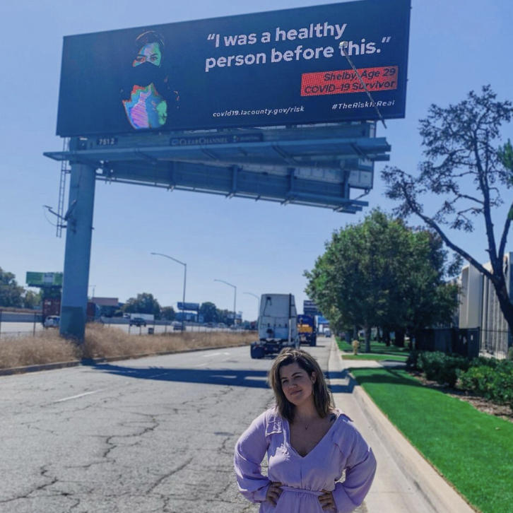 Shelby Hedgecock stands in front of a billboard from a Los Angeles County public health campaign that features her as a long COVID patient.