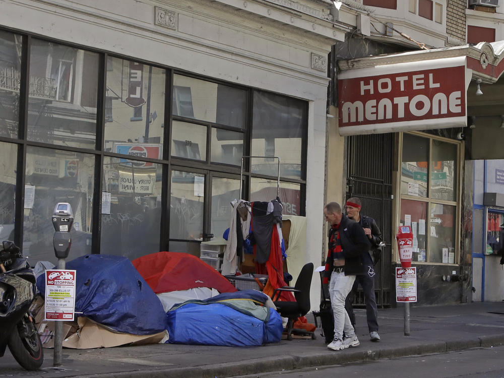 Pedestrians walk along the edge of a sidewalk to avoid tents and sleeping bags, on April 13, 2020, in the tenderloin area of San Francisco.