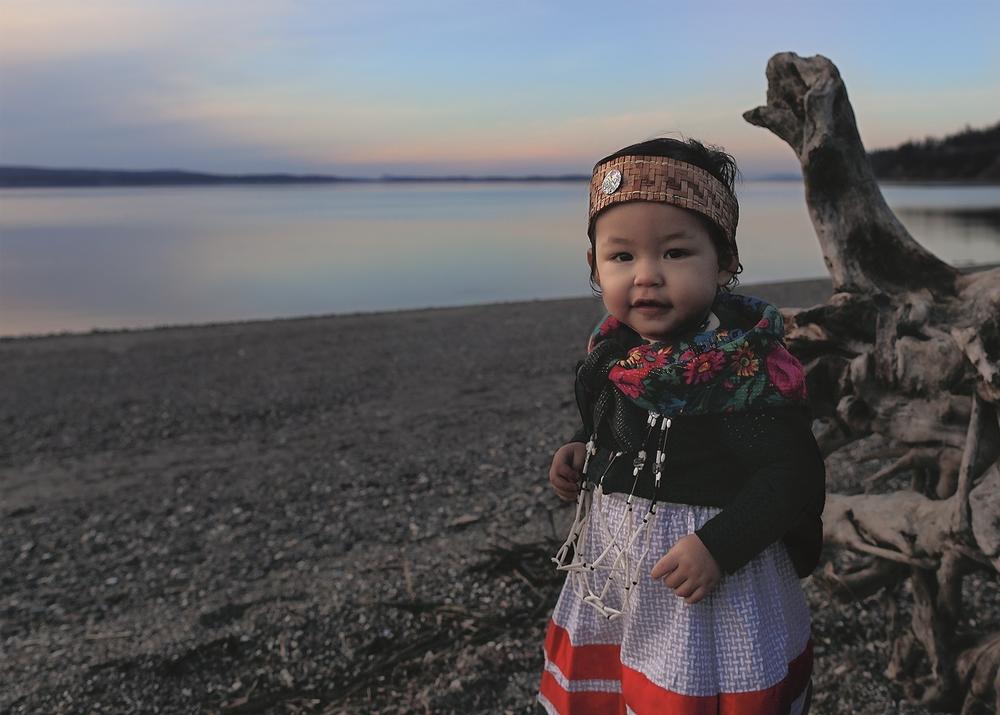 Matika Wilbur dedicates <em>Project 562</em> to her daughter Alma Bee, who was one year old when this photo was taken. Alma Bee is standing along the coast of Washington State on traditional homelands of the Tulalip tribe, with Swinomish land in the background, both reflecting her ancestry.