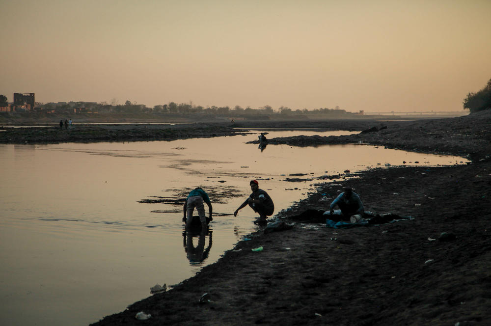 Men wash and sift through industrial ashes in Ravi River water in a large pan, picking out bits of copper and steel that they resell to metal recyclers. The men say they can make a decent living this way — a few dollars a day.