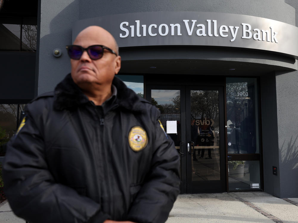 A security guard at Silicon Valley Bank monitors a line of people outside the office in Santa Clara, Calif., on March 13, 2023. The Fed admitted it was partly to blame for the collapse of the lender in a scathing report on Friday.
