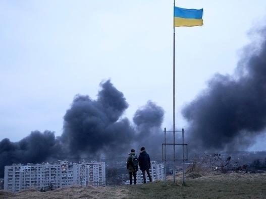 People standing near a Ukrainian national flag watch as dark smoke billows following an air strike in the western Ukrainian city of Lviv, on March 26, 2022.