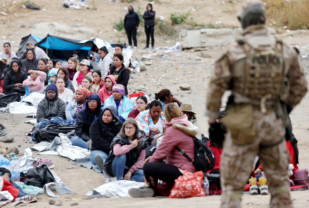 Migrants in a makeshift camp between U.S. and Mexico border walls sit as a Customs and Border Protection officer keeps watch.