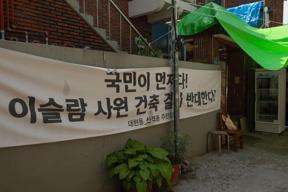 A banner hung in the alleyway opposes the mosque's construction, and says that Korean citizens' interests should be come first. At right, a replica pig's head sits on a chair, and next to it, a few feet from the Islamic center sits a refrigerator with three pig heads in it.