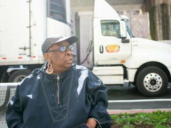 Community activist Margaret Gordon sits on a bench in West Oakland with the BART tracks behind her on March 4, 2022, as a semi-truck stops on 7th Street, on a popular trucking route to the nearby Port of Oakland.
