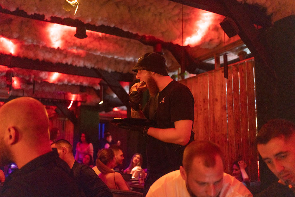 A man eats a burger as he walks to his table, wearing gloves to keep his hands clean.