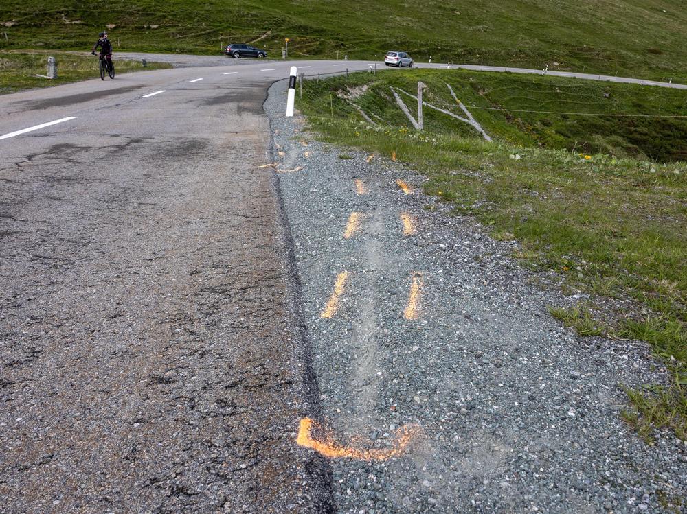 Paint outlines of what look to be two tire tracks leading off the road at the curve where Swiss rider Gino Mäder fell during a high-speed descent of the fifth stage of the Tour de Suisse. Mäder, 26, died from injuries he sustained when he plunged into a ravine.
