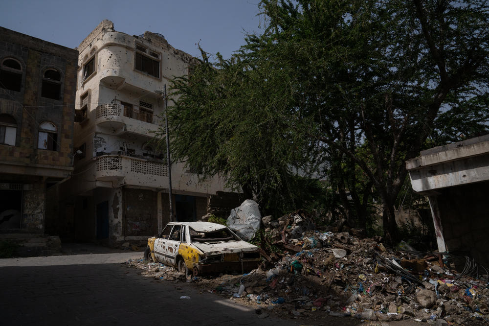 A car and building are riddled with bullet holes in Al Dawah neighborhood. And trash piles up around the neglected car. Services in this area are non-existent though people continue to live here.