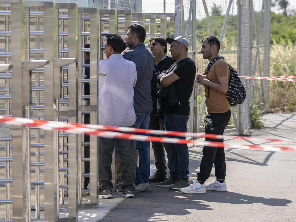 A group of men wait at the turnstiles and speak with survivors, mostly from Pakistan, of a deadly migrant boat sinking at a migrant camp in Malakasa north of Athens, on Monday, June 19, 2023.