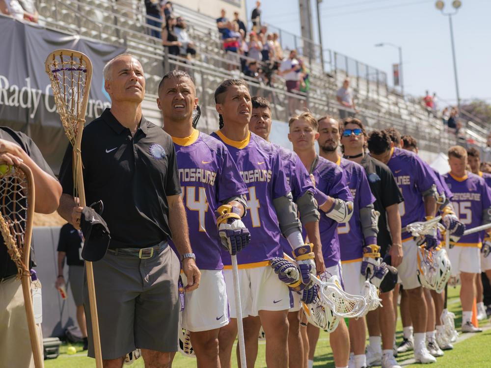 As the Haudenosaunee Nationals listen to their national anthem, Head Coach Lars Tiffany (left) holds a traditionally-crafted lacrosse stick handmade by Alfie Jacques, a renowned stick maker who recently died.