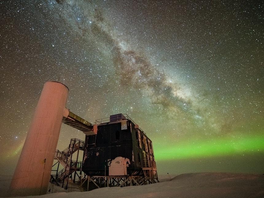 The IceCube Lab under a starry, night sky, with the Milky Way appearing over low auroras in the background.