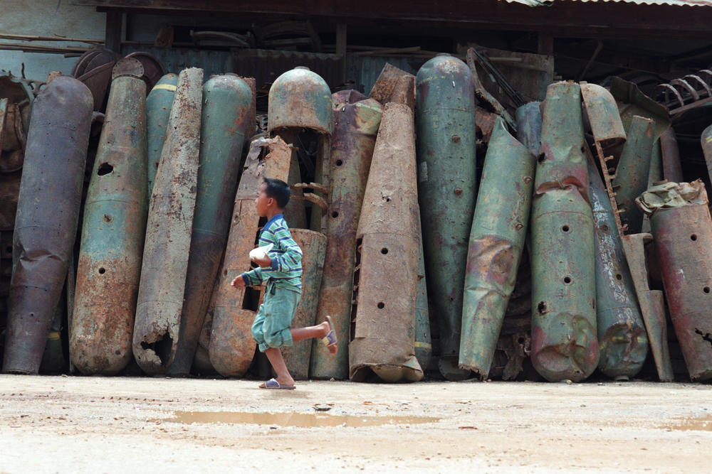 A boy runs past rusted halves of cluster bomb shells and other military hardware in a scrap metal shop along the main street in Phonsavanh, Laos on August 1, 1998.
