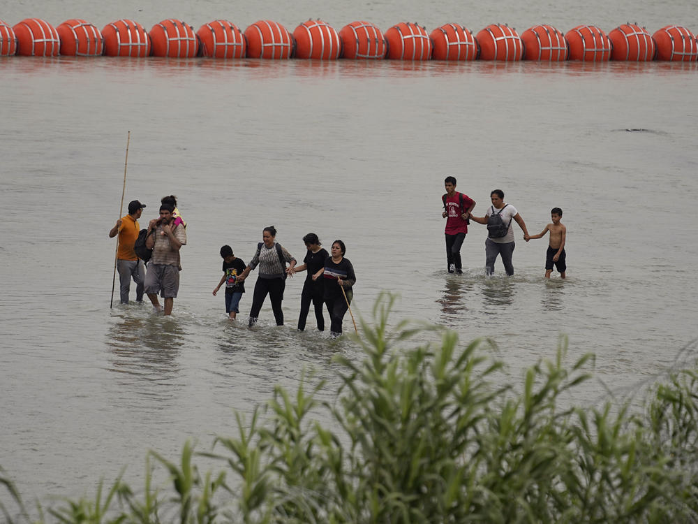 Migrants who crossed the Rio Grande from Mexico walk past large buoys being deployed as a border barrier on the river in Eagle Pass, Texas, on Wednesday.