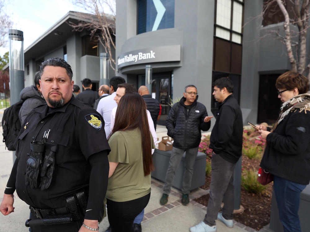 A security guard monitors a line of people outside of a Silicon Valley Bank office in Santa Clara, Calif., on March 13, 2023. The collapse of SVB sparked a period of intense volatility in markets as investors feared about the health of the smaller lenders.
