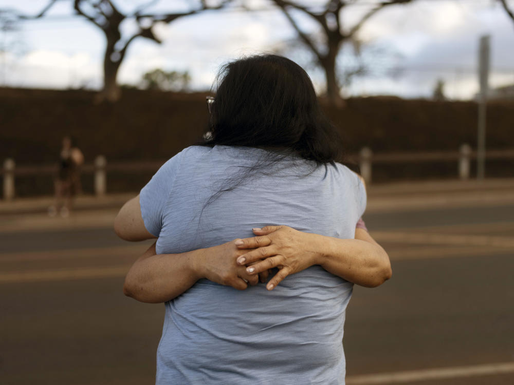 Carelle Calvan in blue T-shirt, hugs Nora Bulosan as they gather near a checkpoint in hopes to get access to their home destroyed in recent wildfires in Lahaina, Hawaii, Wednesday, Aug. 16, 2023.
