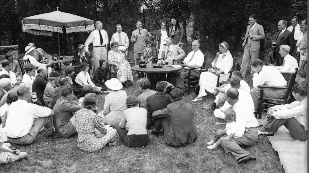 President Franklin Roosevelt reads to his guests as he and first lady Eleanor Roosevelt, at table, host a Labor Day picnic at their residence in Hyde Park, N.Y., Sept. 3, 1934.