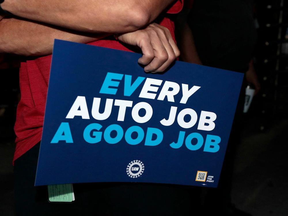 UAW supporters and members hold signs outside the Stellantis Assembly Plant in Sterling Heights, Mich., on July 12. Electric vehicles may not be at the heart of the contract talks between the UAW and the Detroit automakers, but as the sign subtly shows, they loom large in the negotiations.