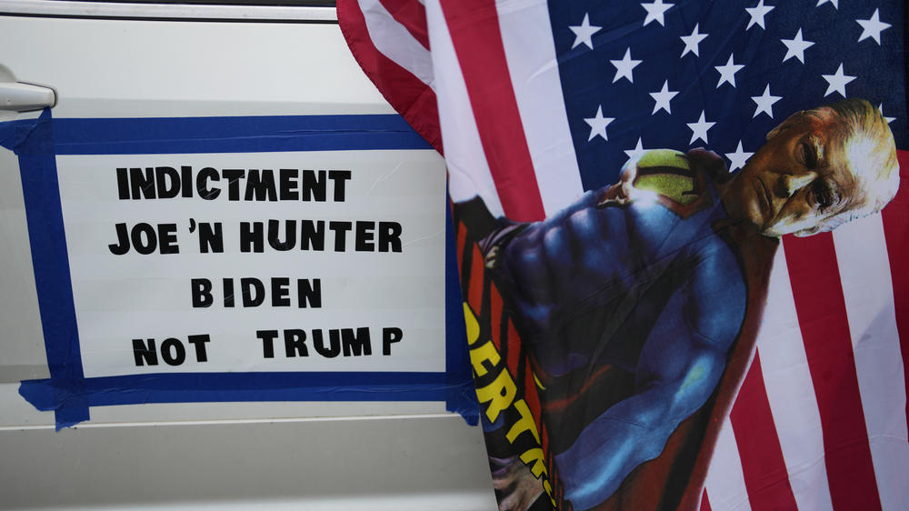 A flag depicting former President Donald Trump as a superhero hangs from a car with a sign calling for President Biden and his son Hunter Biden to be indicted instead of Trump, as Trump supporters gather at Tropical Park in Miami on June 11.