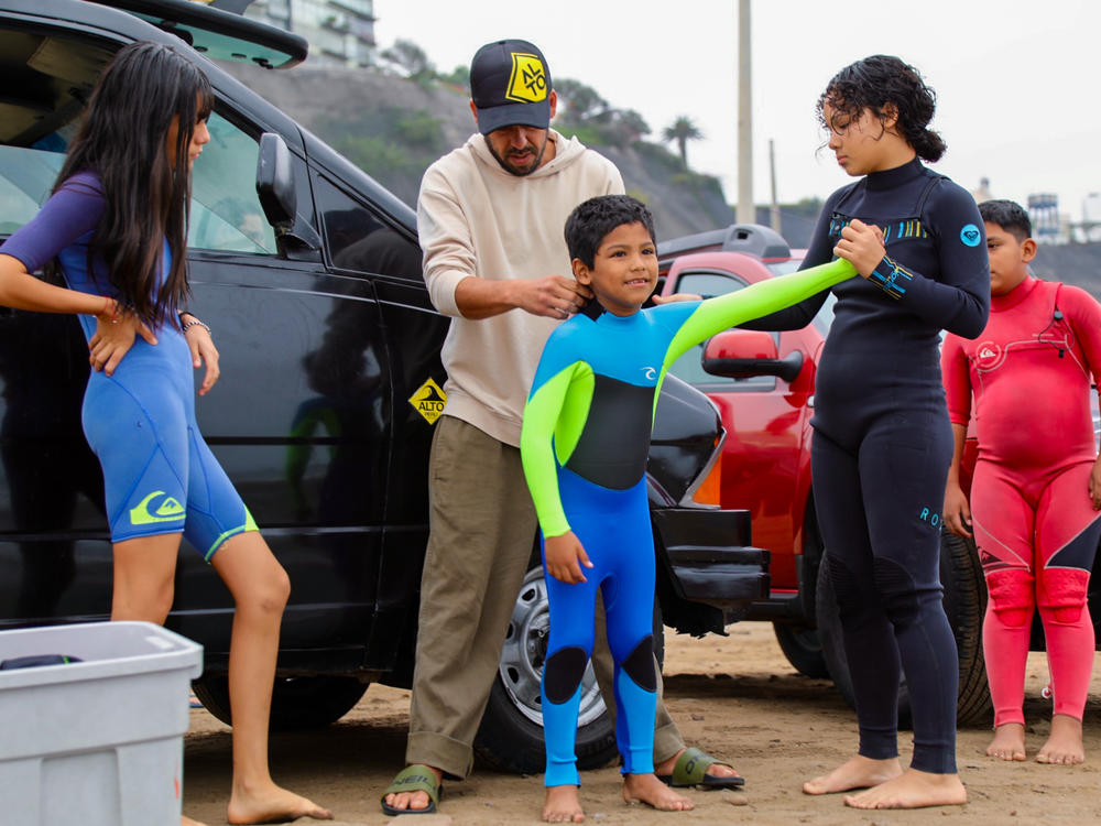 Boran Bumovich Hignio, a 7-year-old surfer, gets help with his wetsuit from Diego Villarán, who founded the local surf school, and other staffers.