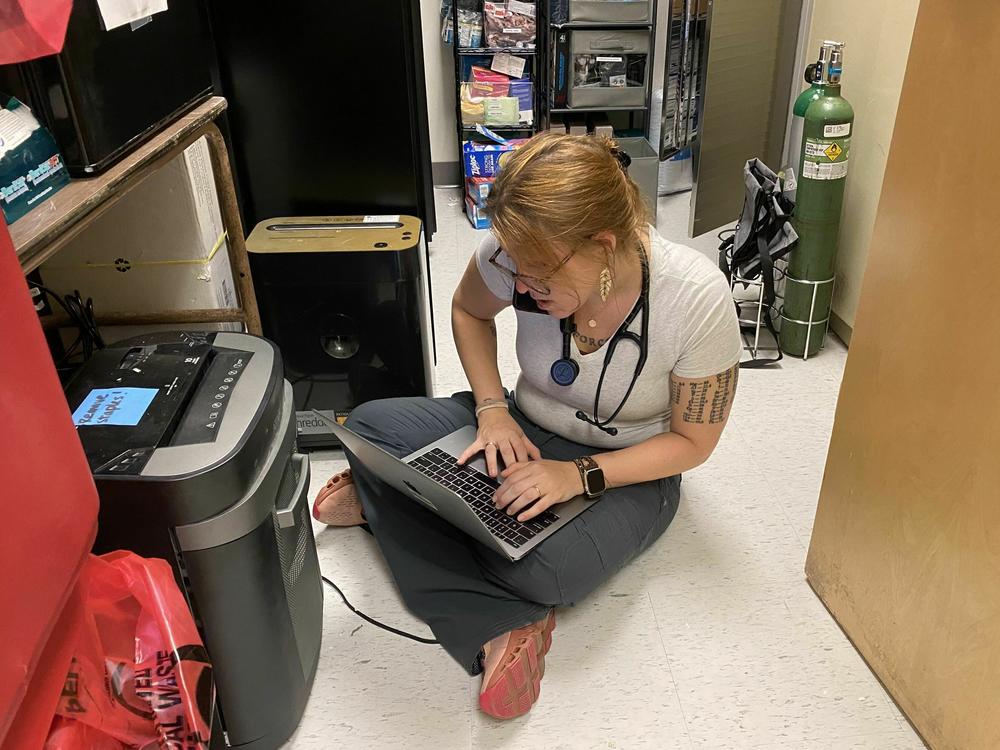 Elyse Stevens, a primary care doctor who specializes in addiction, sits in a closet at a shelter, where she delivers care. She's on the phone with a pharmacy, while simultaneously printing out papers for a patient.
