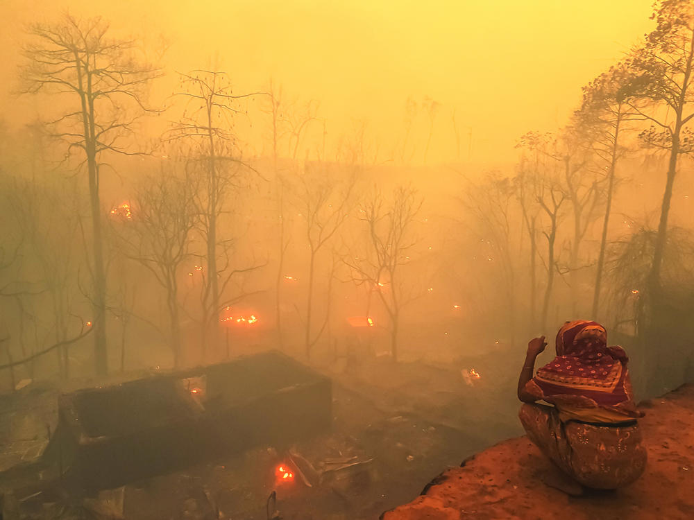 The fire that tore through the Bangladesh refugee camp in March 2021 killed 15 people and displaced some 45,000. Pictured, a woman looks on at her ruined shelter while fire still rages.