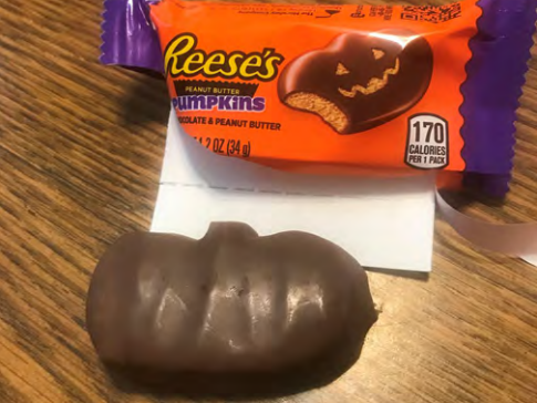 A Reese's Peanut Butter Pumpkin with no carved-out face next to the open wrapper with a jack-o'-lantern design.
