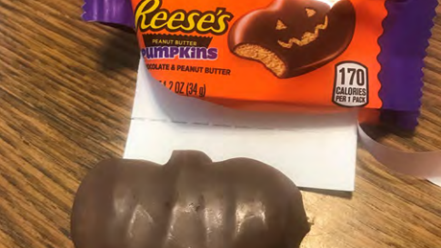 A Reese's Peanut Butter Pumpkin with no carved-out face next to the open wrapper with a jack-o'-lantern design.
