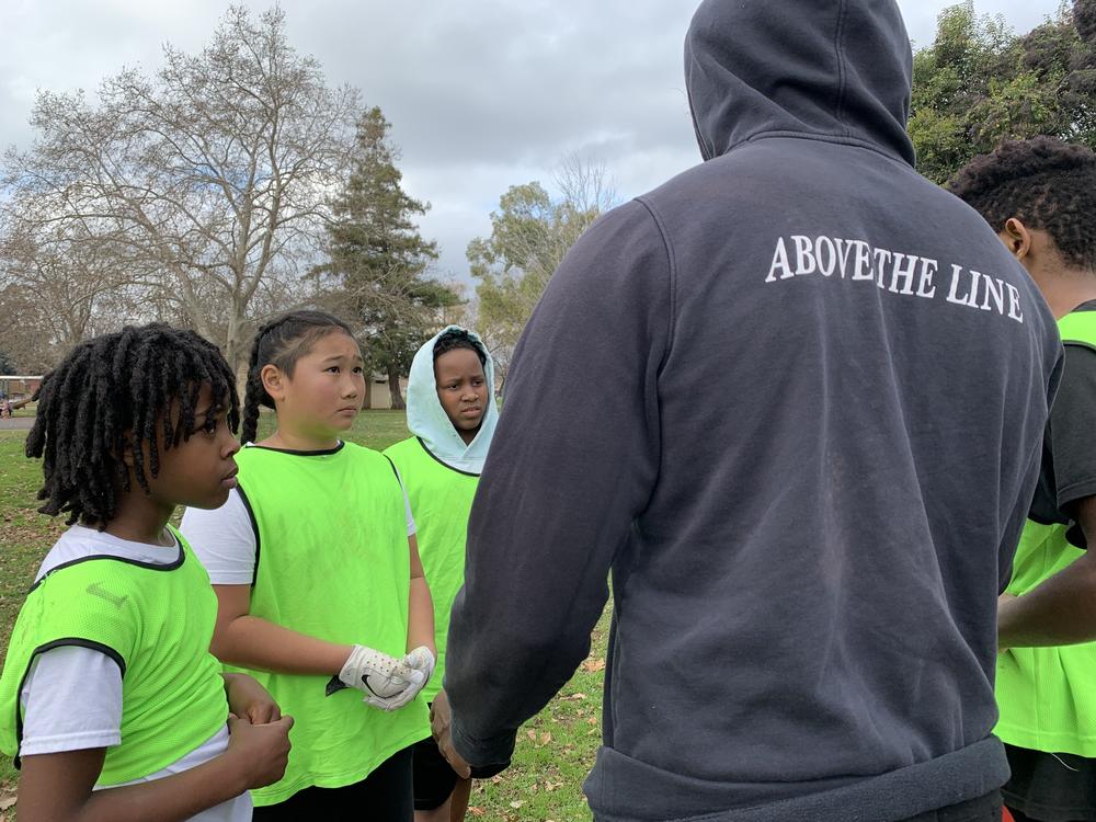 Coach Damaree Morris, holding a team meeting with Monte Johnson, Kainoa Navarrete, and King Simon, says parents should have a choice as to whether or not they want their kids to participate in tackle football.