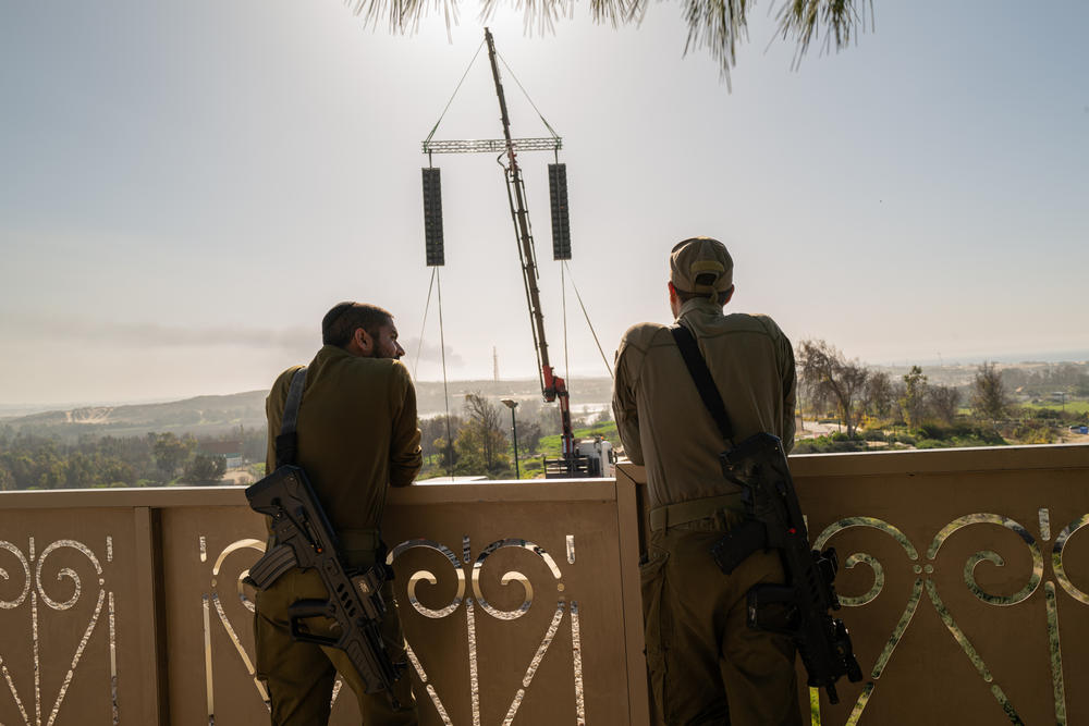 Israeli soldiers listen as the concert is broadcast over giant speakers in the hopes that Alon Ohel can hear it. In the distance, smoke can be seen rising from Gaza.