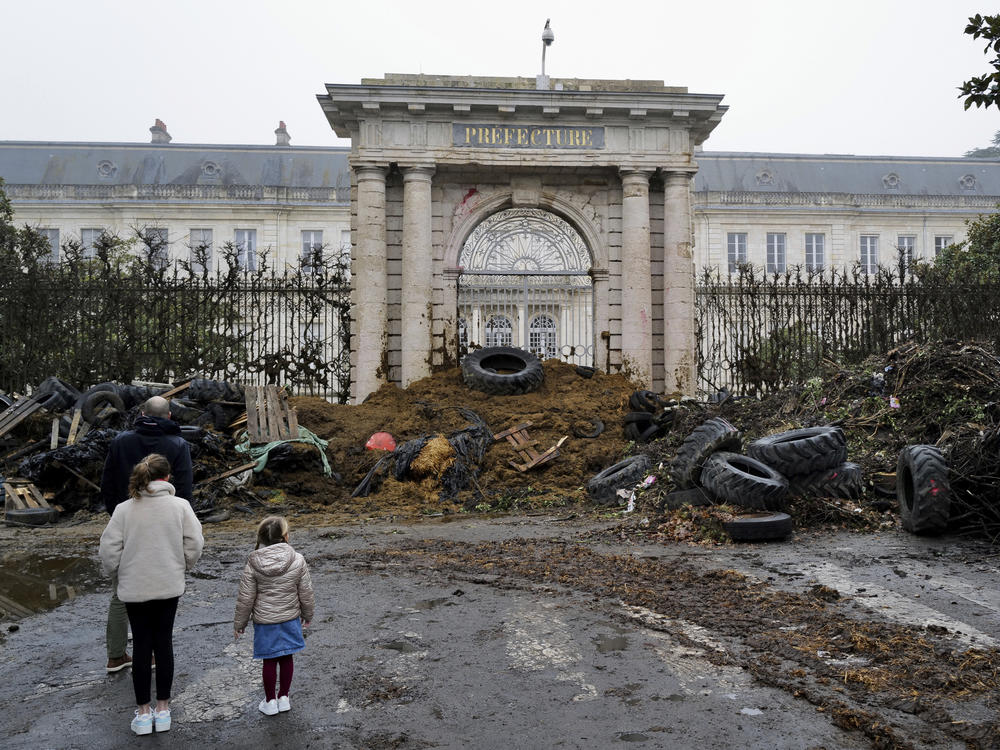 People watch slurry, manure and tyres dumped by farmers at the entrance of the local state administration building, in Agen, southwestern France, Saturday, Jan. 27, 2024. French farmers have vowed to continue protesting and are maintaining traffic barricades on some of the country's major roads.
