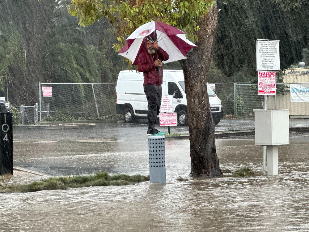 A man carrying an umbrella stands perched above a flooded street in Ventura, Calif., Sunday, Feb. 4, 2024.
