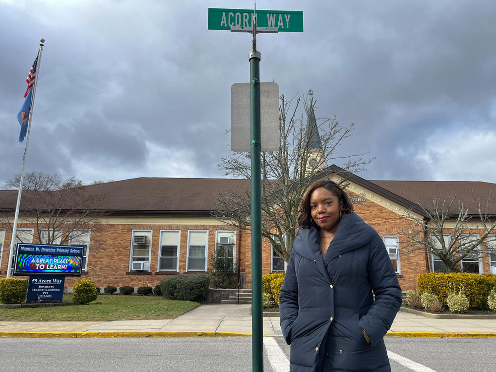 Lisa Ortiz, a resident of Lakeview, N.Y., stands under the new street sign for Acorn Way in the predominantly white village of Malverne, where many Black students from Lakeview attend schools. The street was previously named Lindner Place after a leader of the Ku Klux Klan in New York.