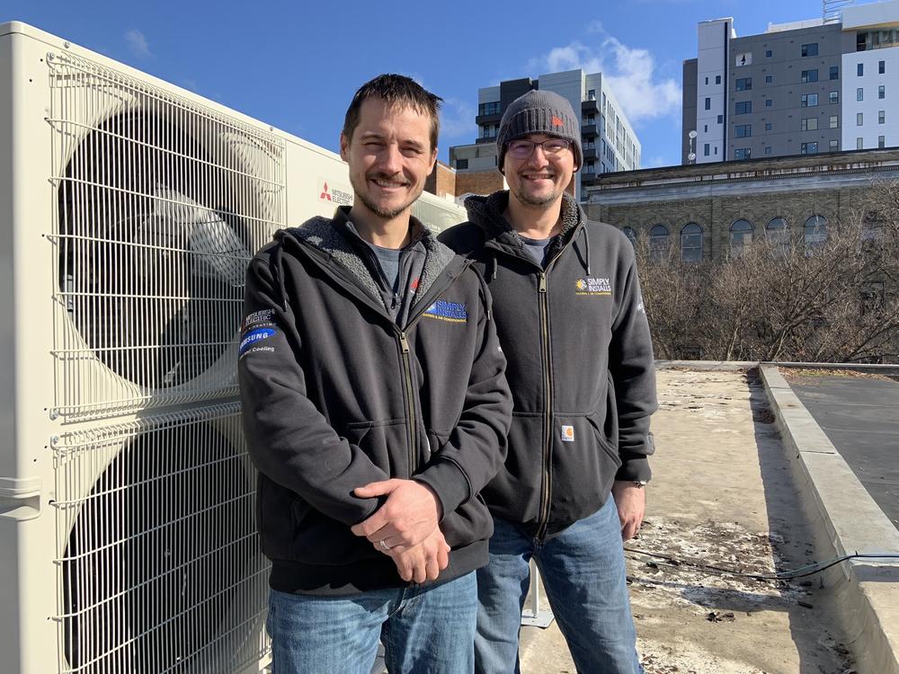 Brothers Logan and AJ Cole, owners of Simply Installs Heating & Air Conditioning, stand next to a heat pump they installed at Ithaca Piercing & Tattoo. This project was part of a citywide program to convert gas furnaces in buildings to climate friendly electric.