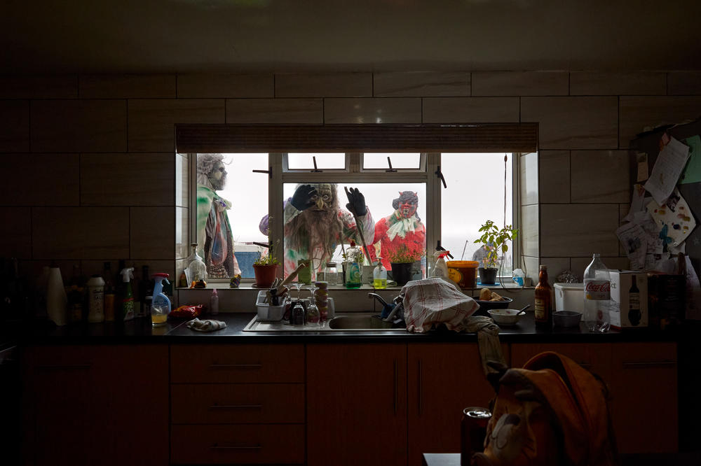 Several Okalolies peer into the kitchen of a house belonging to one of their own. Although they are careful not to frighten children and the elderly too much, they are expected to make light mischief, and will attempt to soak any woman they find with a garden hose.