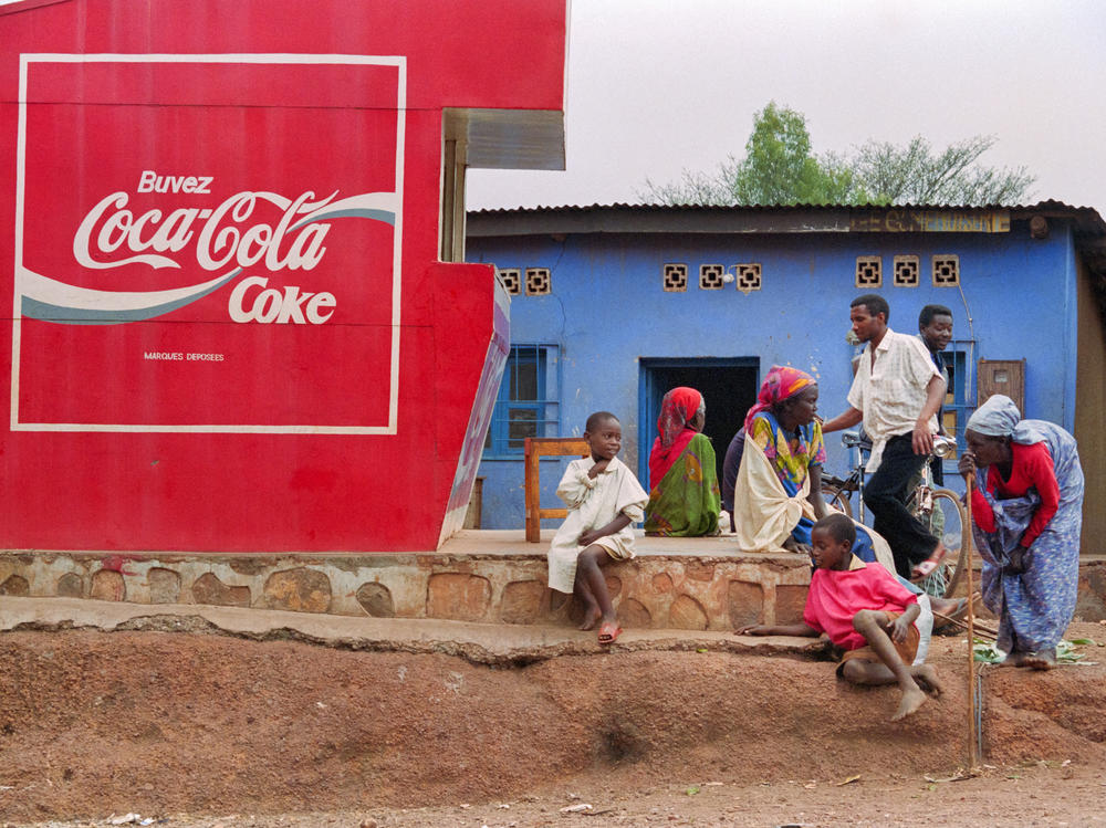 A group of Rwandans waits for transportation near a Coca Cola placard. The photo is from 1994.