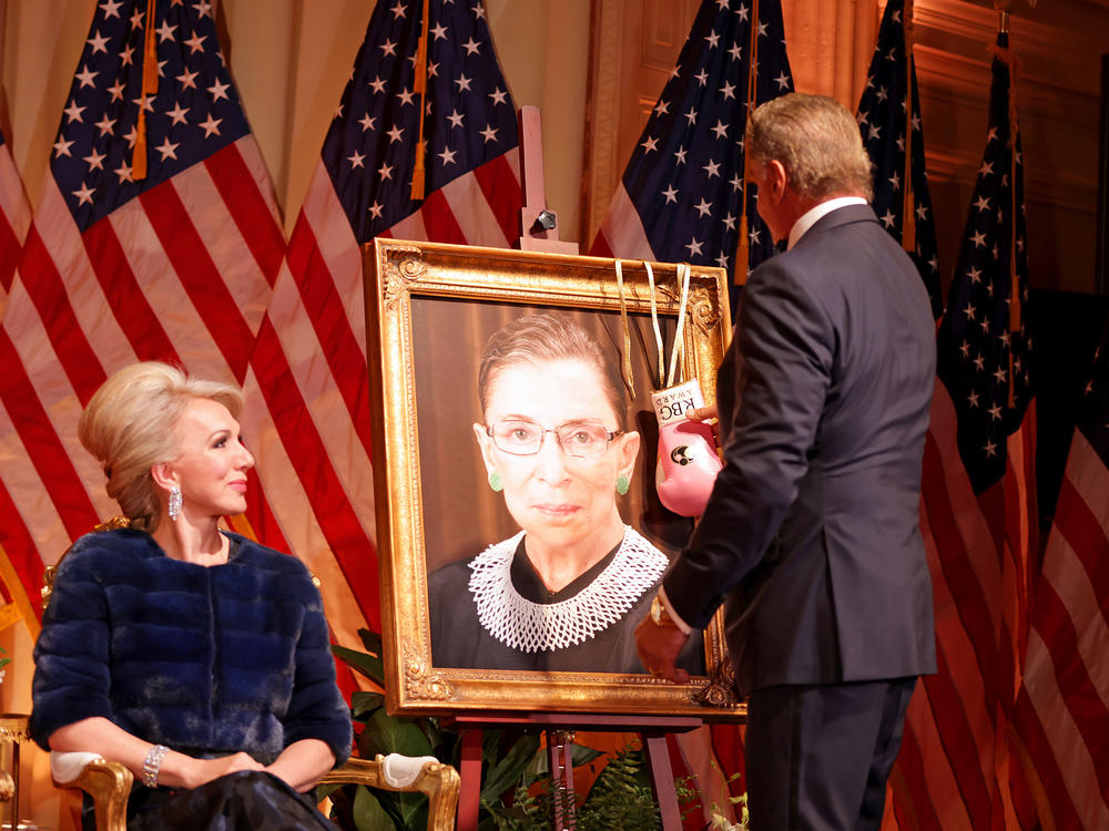 Sylvester Stallone hangs a pair of pink boxing gloves on a portrait of the late Supreme Court Justice Ruth Bader Ginsburg at the Justice Ruth Bader Ginsburg Woman of Leadership Award in 2022, as Julie Opperman, chair of the Dwight D. Opperman Foundation, looks on.