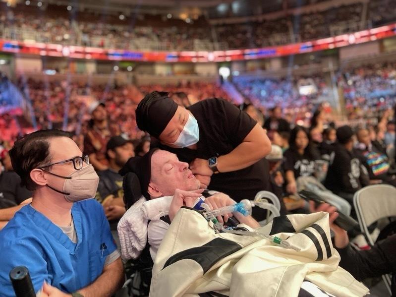 Kevin Carlson, with nurse Joshua Lee (right) and respiratory therapist Eric Mathewson (left), watches a WWE match on October 2, 2023 in San Jose, Calif.