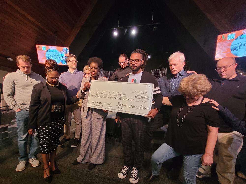 Members of the Sycamore Creek Church congregation bow their heads and pray over Justice League of Greater Lansing leaders, Willye Bryan and Prince Solace, as they hold a large check.