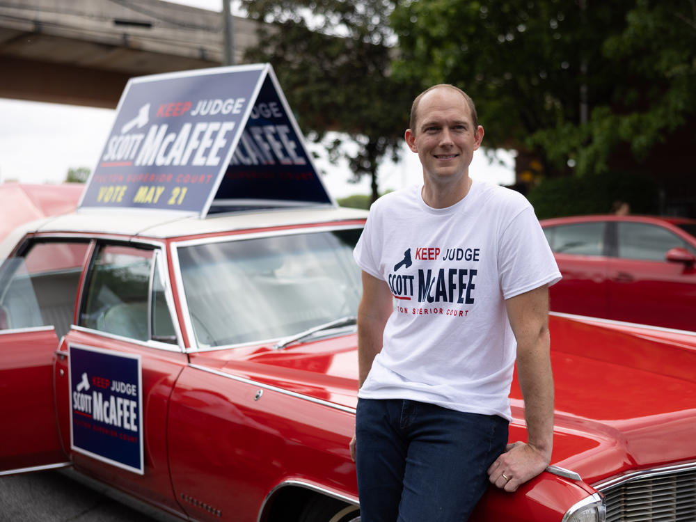 Fulton Superior Court Judge Scott McAfee leans against a 1965 Cadillac Fleetwood that has been in his family for years, while campaigning at a parade in Atlanta on April 27. McAfee, the trial judge in the Georgia election interference case, is up for reelection this month in a nonpartisan race.